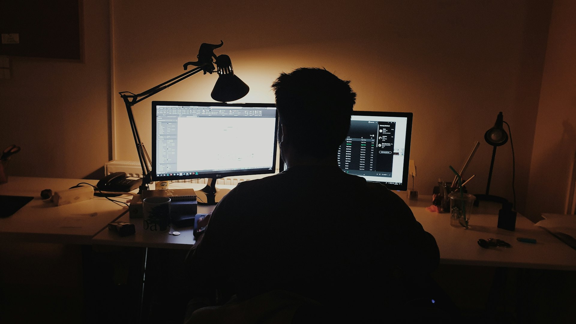 Persona de espaldas trabajando frente a dos monitores en una oficina en penumbra